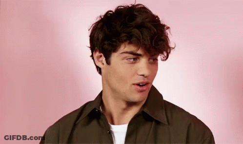 A Young Man With Curly Hair Is Standing In Front Of A Pink Background And Looking At The Camera . GIF