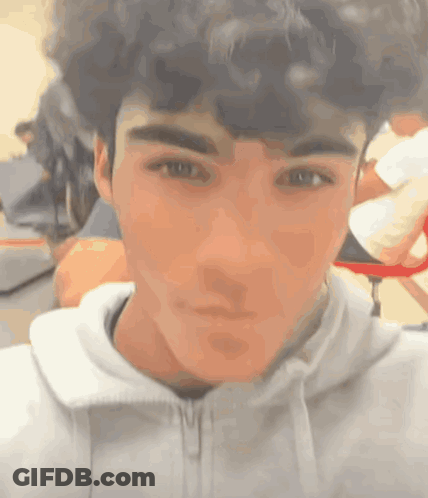 A Young Man With Curly Hair Is Taking A Selfie In A Classroom . GIF