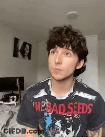 A Young Man With Curly Hair Is Wearing A Bad Seeds T-Shirt And Looking At The Camera . GIF