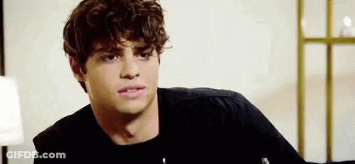 A Young Man With Curly Hair Is Wearing A Black Shirt And Sitting In A Room . GIF