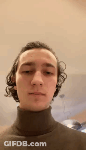 A Young Man With Curly Hair Is Wearing A Black Turtleneck Sweater And Looking At The Camera . GIF