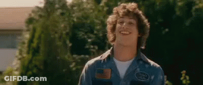 A Young Man With Curly Hair Is Wearing A Blue Shirt And Smiling In Front Of Trees . GIF