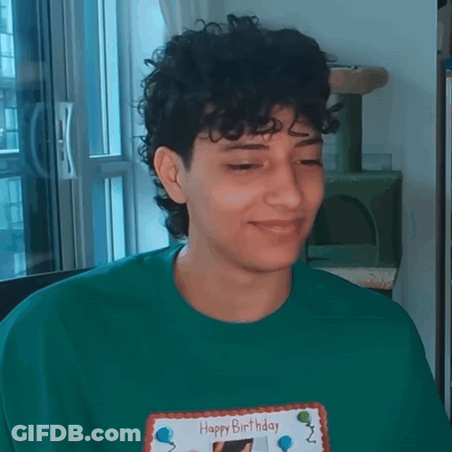 A Young Man With Curly Hair Is Wearing A Green Shirt And Holding A Happy Birthday Card . GIF