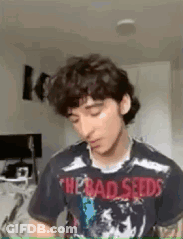 A Young Man With Curly Hair Is Wearing A T-Shirt With The Bad Seeds On It . GIF