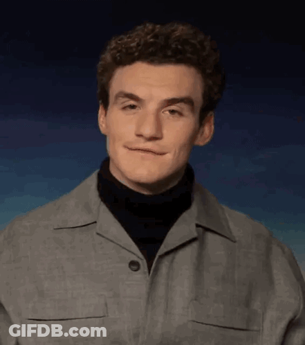 A Young Man With Curly Hair Is Wearing A Turtleneck And A Grey Jacket . GIF