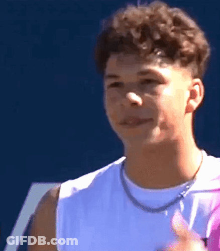 A Young Man With Curly Hair Wearing A White Tank Top And A Necklace Is Smiling . GIF