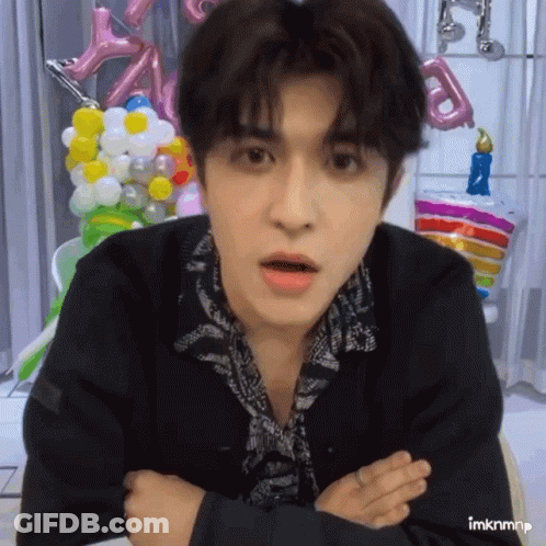 A Young Man With His Arms Crossed Is Looking At The Camera With Balloons In The Background GIF