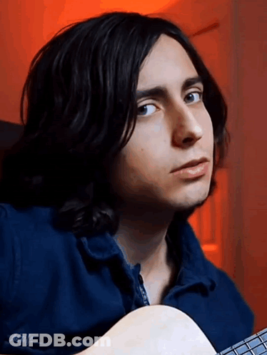 A Young Man With Long Black Hair Is Holding A Guitar And Wearing A Blue Jacket GIF