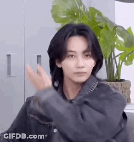 A Young Man With Long Black Hair Is Standing In Front Of A Potted Plant In A Room . GIF
