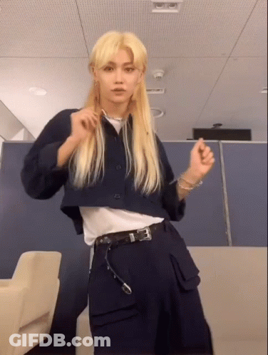 A Young Man With Long Blonde Hair Is Standing In A Room Wearing A Blue Jacket And Black Pants . GIF