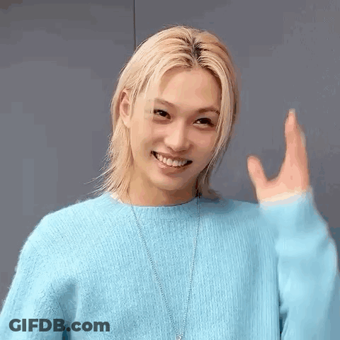 A Young Man With Long Blonde Hair Is Wearing A Blue Sweater And Necklace . GIF