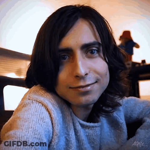 A Young Man With Long Dark Hair Is Wearing A Sweater And Smiling . GIF