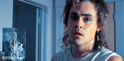 A Young Man With Long Hair And Earrings Is Standing In Front Of A Door . GIF