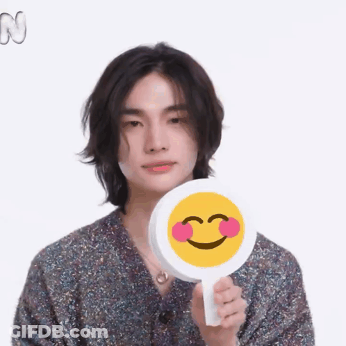 A Young Man With Long Hair Is Holding A Smiley Face With Pink Cheeks . GIF