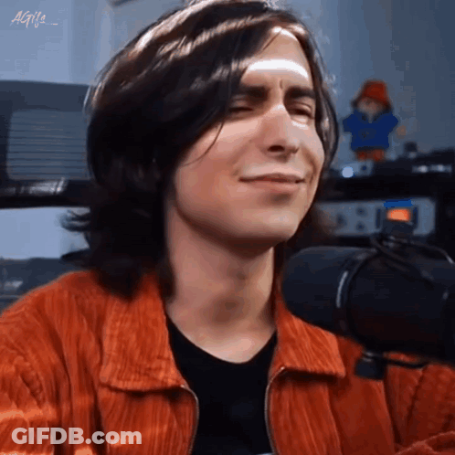 A Young Man With Long Hair Is Smiling In Front Of A Microphone . GIF