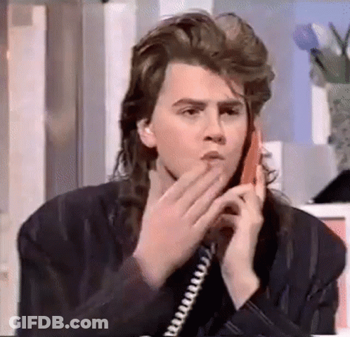 A Young Man With Long Hair Is Talking On A Telephone . GIF