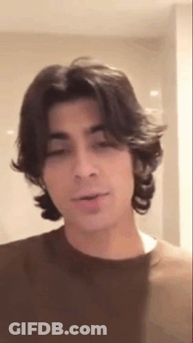 A Young Man With Long Hair Is Wearing A Brown Shirt And Talking . GIF