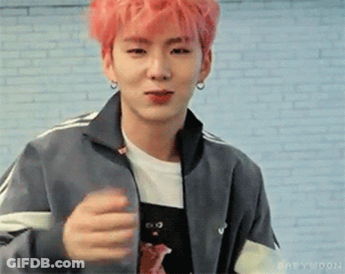 A Young Man With Pink Hair And A Gray Jacket Is Standing In Front Of A White Brick Wall . GIF