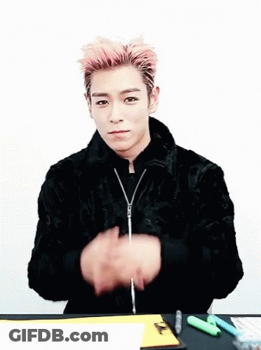 A Young Man With Pink Hair Is Sitting At A Table Making A Heart With His Hands GIF