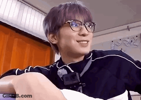A Young Man With Purple Hair And Glasses Is Sitting On A Couch . GIF