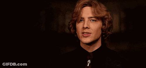 A Young Man With Red Hair Is Wearing A Black Shirt And Tie And Looking At The Camera . GIF