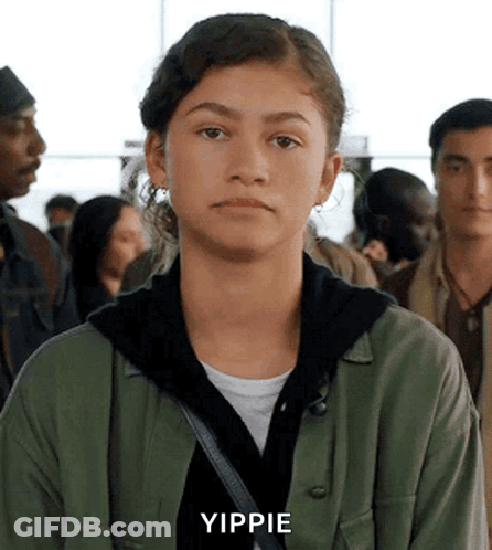 A Young Woman In A Green Jacket Is Standing In A Crowd Of People And Making A Funny Face . GIF