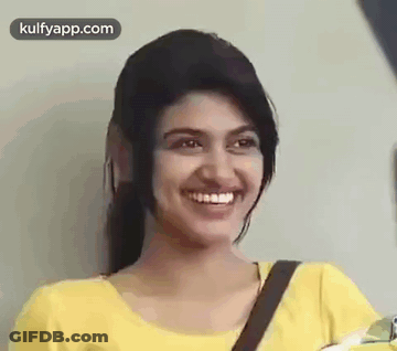 A Young Woman In A Yellow Shirt Is Smiling And Looking At The Camera . GIF