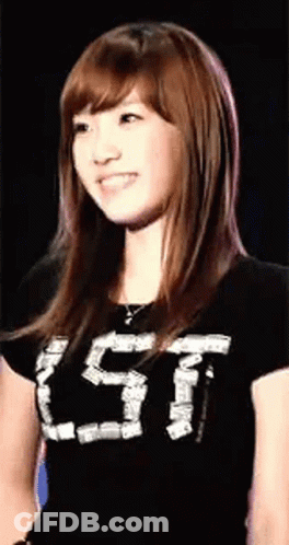 A Young Woman Wearing A Black T-Shirt With The Letter T On It Is Smiling And Looking At The Camera . GIF