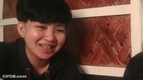 A Young Woman With Braces On Her Teeth Is Smiling While Sitting In Front Of A Wooden Wall . GIF