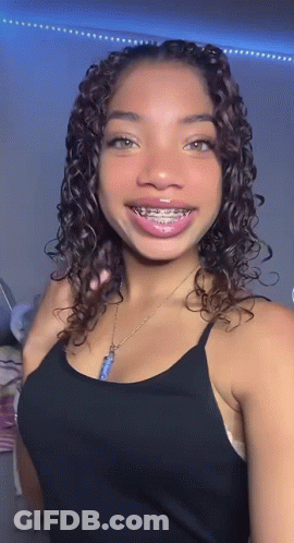 A Young Woman With Curly Hair And Braces On Her Teeth Is Wearing A Black Tank Top And A Necklace . GIF