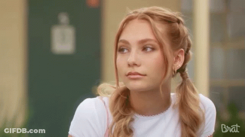 A Young Woman With Pigtails And Earrings Is Sitting In Front Of A Building . GIF