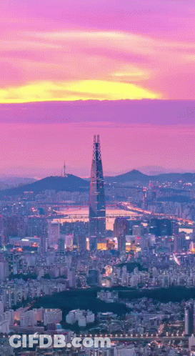 An Aerial View Of A City At Sunset With A Tall Building In The Foreground GIF