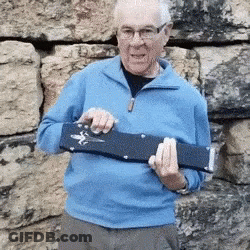 An Elderly Man In A Blue Sweater Is Holding A Guitar Case In Front Of A Stone Wall . GIF