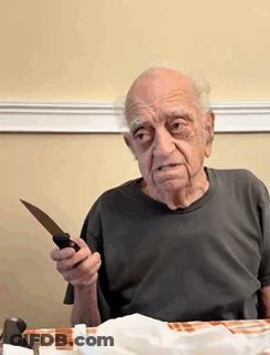 An Elderly Man Is Sitting At A Table Holding A Knife In His Hand GIF