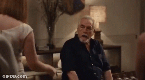 An Elderly Man Is Sitting On A Couch In A Living Room With A Woman Standing Next To Him . GIF