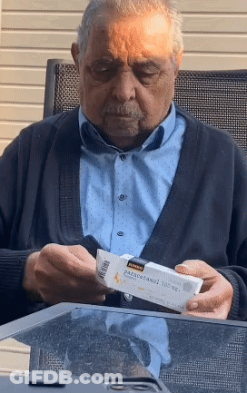 An Elderly Man Sitting At A Table Looking At A Box Of Paracetamol GIF