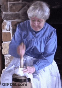 An Elderly Woman In A Blue Shirt And White Apron Is Using A Wooden Hammer GIF
