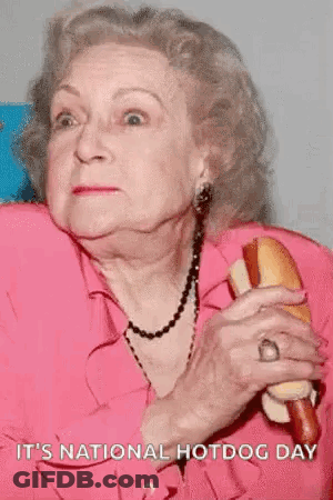An Elderly Woman In A Pink Shirt Is Holding A Hot Dog And Says It 'S National Hotdog Day . GIF