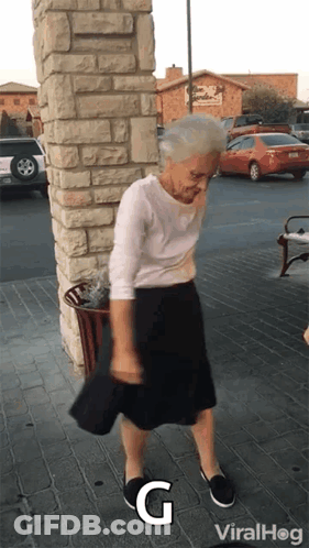An Elderly Woman Is Dancing In Front Of A Brick Building With The Letters G On The Ground GIF