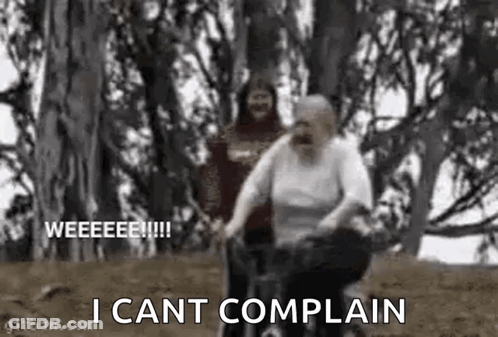 An Elderly Woman Is Riding A Bike In The Woods While A Man Looks On . GIF