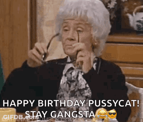 An Elderly Woman Is Sitting At A Table Holding Glasses And Saying Happy Birthday Pussycat Stay Gangsta GIF