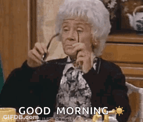 An Elderly Woman Is Sitting At A Table Holding Glasses And Says Good Morning GIF