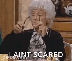 An Elderly Woman Is Sitting At A Table Holding Glasses And Says `` I Ain T Scared '' . GIF