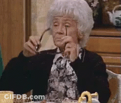 An Elderly Woman Is Sitting At A Table Holding Glasses In Her Hand . GIF