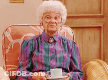 An Elderly Woman Is Sitting In A Chair Holding A Cup And Saucer GIF