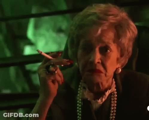 An Elderly Woman Is Smoking A Cigarette And Waving At The Camera In Front Of A Green Background . GIF