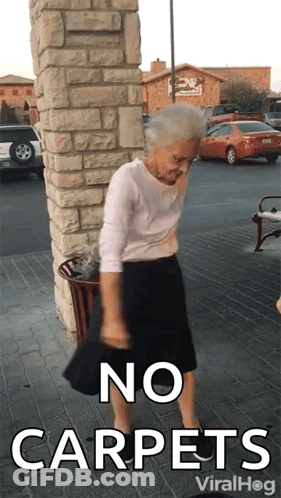 An Elderly Woman Is Standing In Front Of A Brick Wall With The Words No Carpets Written On It GIF