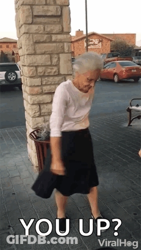 An Elderly Woman Is Standing In Front Of A Brick Wall With The Words " You Up " On The Bottom GIF