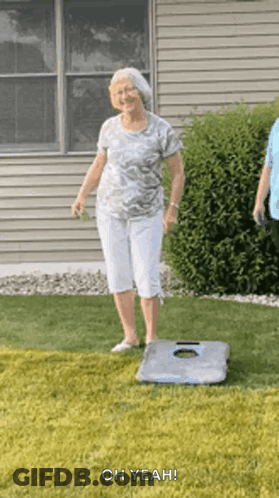 An Elderly Woman Is Standing On A Cornhole Board In A Grassy Yard . GIF