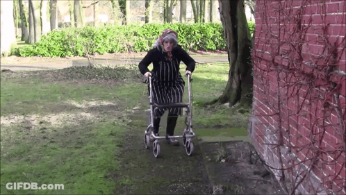 An Elderly Woman Is Walking With A Walker In Front Of A Red Brick Wall GIF